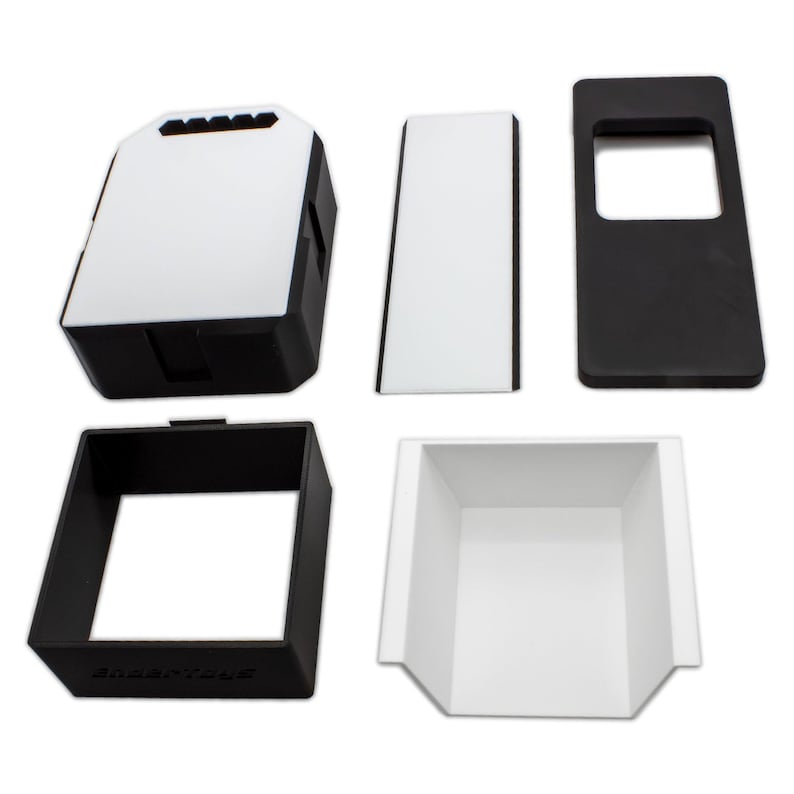 May include: An assortment of black and white plastic components, likely for a larger product. The pieces include a rectangular box with a white top, a rectangular frame, and a tray-like structure. The components are arranged against a white background.