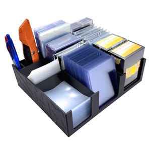May include: A black plastic organizer tray with multiple compartments filled with stacks of clear plastic card sleeves, rigid card holders, two blue pens, and an orange utility knife. The exterior of the tray features a geometric pattern.