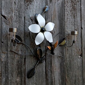 May include: A metal wall decor featuring a white flower with a black center and a hummingbird in flight. The decor is attached to a wooden background and includes two solar lights.