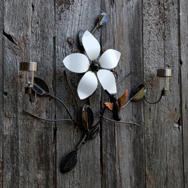 May include: A metal wall decor featuring a white flower with a black center and a hummingbird in flight. The decor is attached to a wooden background and includes two solar lights.