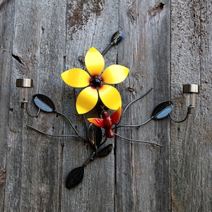 May include: Metal wall art featuring a yellow flower with a red center and a red hummingbird perched on a branch. The art is mounted on a weathered wooden background.