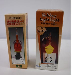 May include: Two boxes of holiday night lights. One box reads "TURKEY BUBBLING NIGHT LIGHT" and features a red and orange turkey-shaped night light. The other box reads "Holiday Night Light With Turkey Topper" and shows a similar night light with a yellow base.