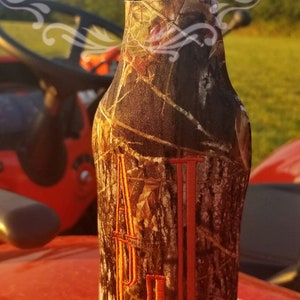 K&ouml;nnte beinhalten: Ein brauner und beigefarbener Camouflage-Bierk&uuml;hler mit dem Buchstaben "J" in Orange auf der Vorderseite bestickt. Der Bierk&uuml;hler befindet sich auf einer roten Oberfl&auml;che.