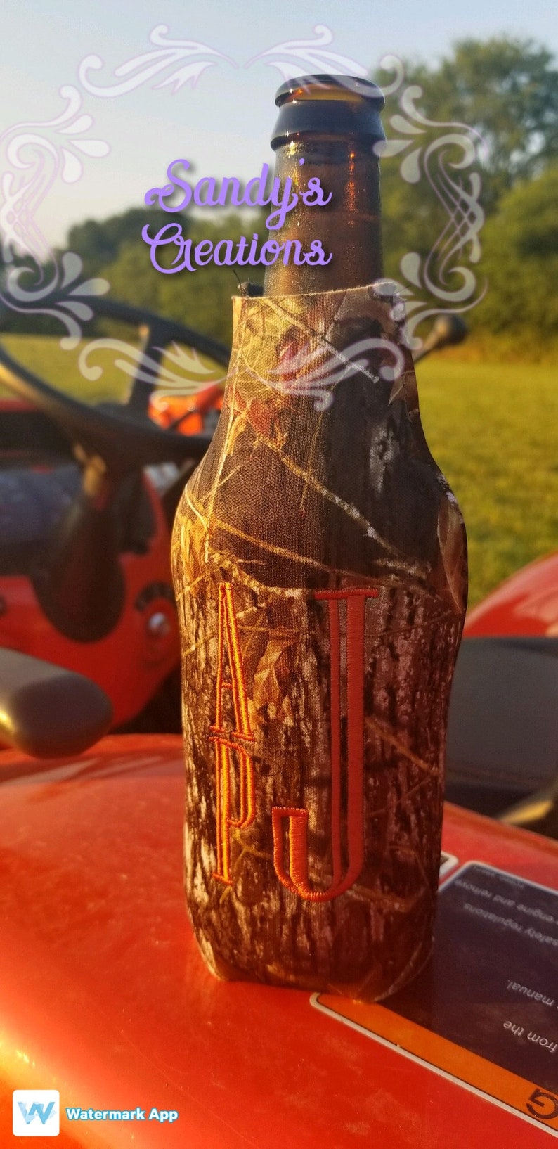 K&ouml;nnte beinhalten: Ein brauner und beigefarbener Camouflage-Bierk&uuml;hler mit dem Buchstaben "J" in Orange auf der Vorderseite bestickt. Der Bierk&uuml;hler befindet sich auf einer roten Oberfl&auml;che.