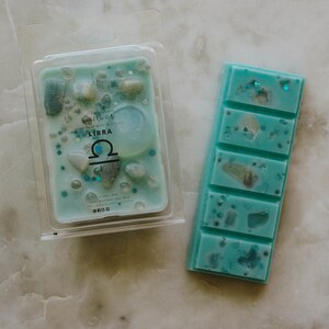 May include: A teal blue wax melt with a Libra zodiac symbol and small white and gold accents. The wax melt is in a clear plastic container with the text "Hand Made with Intention" and "100% Natural Soy Wax".