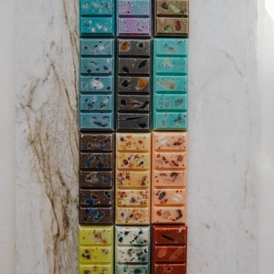 May include: A set of 12 wax melts in various colours and patterns, including blue, green, yellow, orange, brown, and purple. The wax melts are arranged in three rows of four, with each melt having a different design.