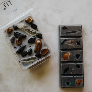 May include: Two wax melts, one in a clear plastic container with a grey wax base and various coloured stones, and the other a grey wax bar with five sections, each with a different coloured stone. The container has the zodiac symbol for Scorpio on it.
