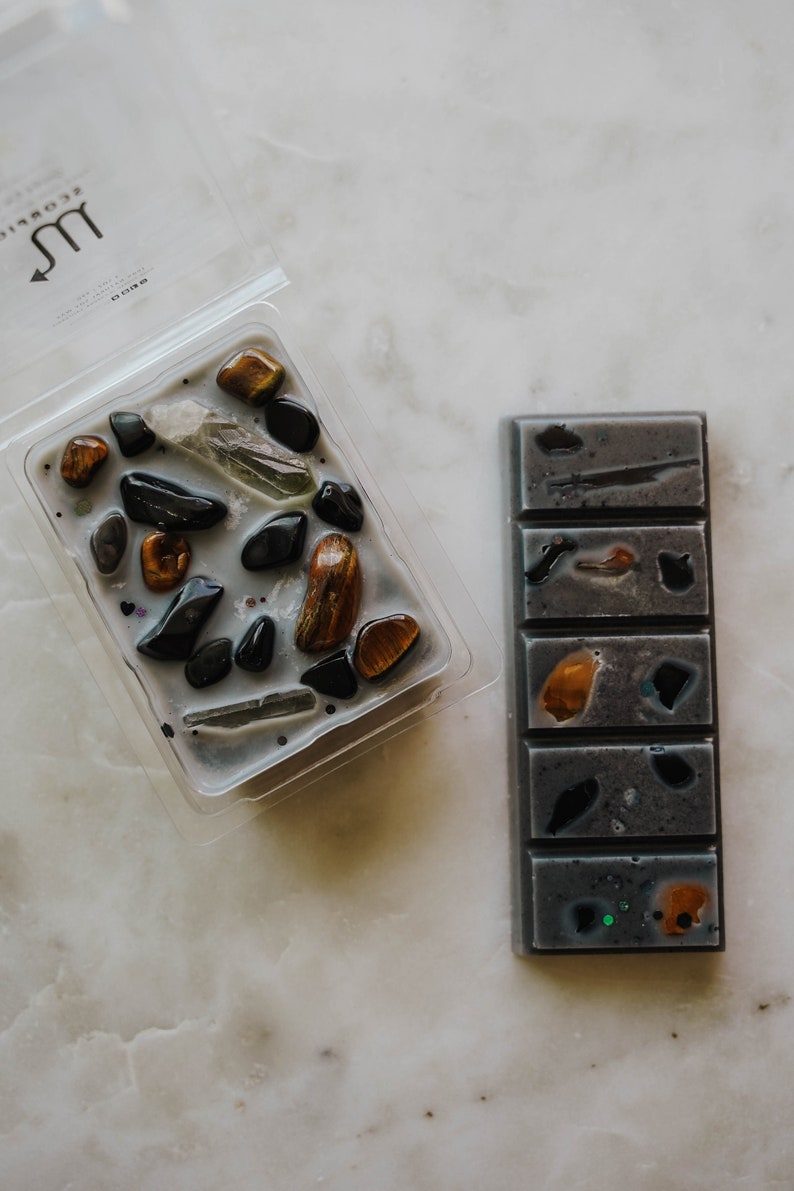 May include: Two wax melts, one in a clear plastic container with a grey wax base and various coloured stones, and the other a grey wax bar with five sections, each with a different coloured stone. The container has the zodiac symbol for Scorpio on it.