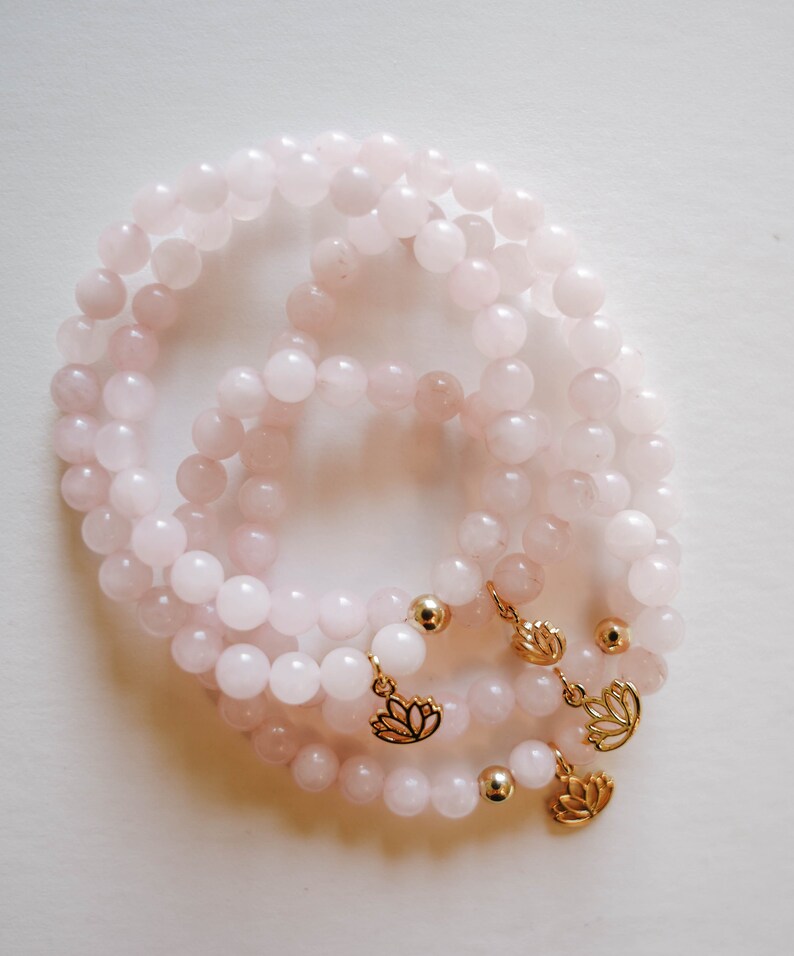 May include: Three pink rose quartz bracelets with gold lotus flower charms. The bracelets are stacked on top of each other.