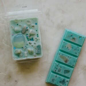 May include: Two wax melts, one in a clear plastic container and one in a bar form. Both are teal blue with white and silver glitter and small white and blue stones. The container has the word "LIBRA" and a Libra zodiac symbol on the top.