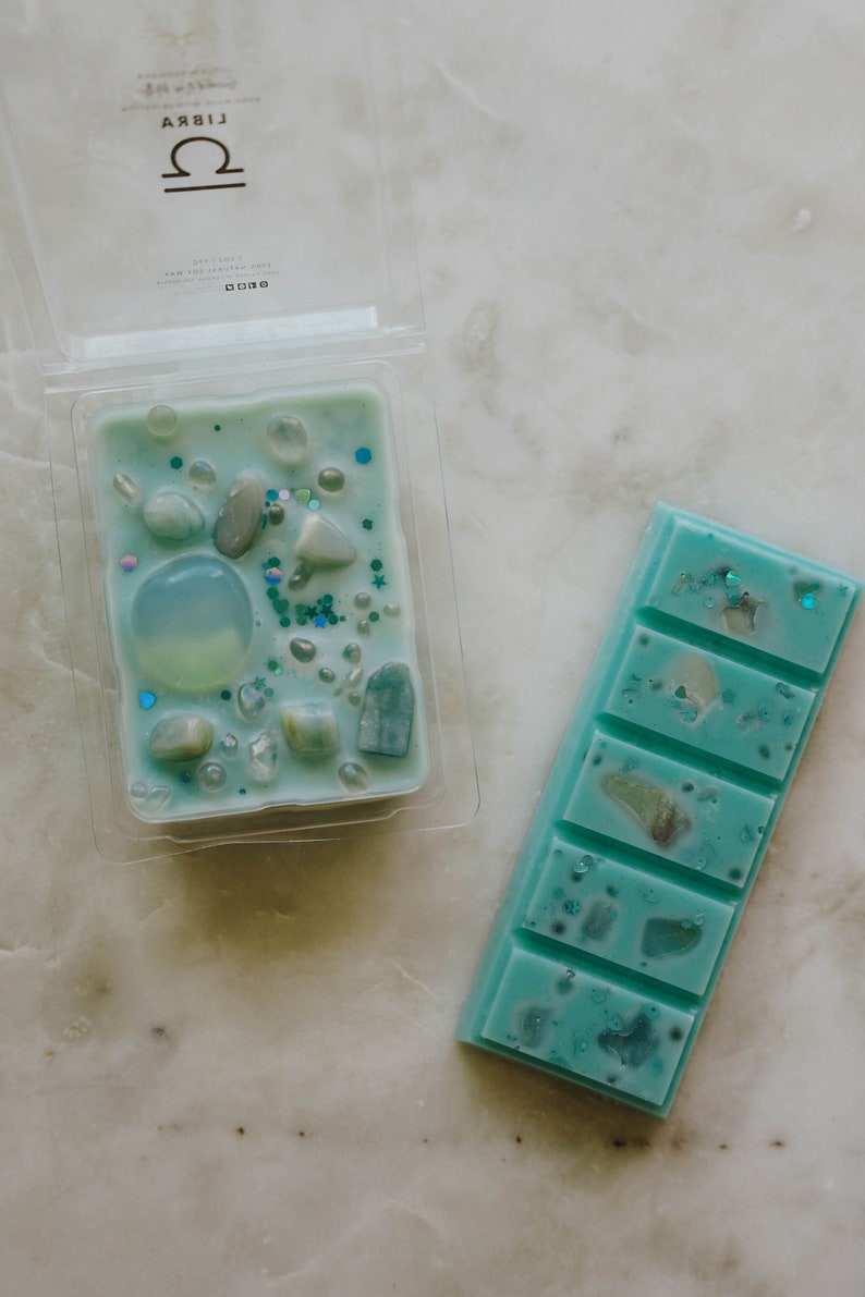 May include: Two wax melts, one in a clear plastic container and one in a bar form. Both are teal blue with white and silver glitter and small white and blue stones. The container has the word "LIBRA" and a Libra zodiac symbol on the top.