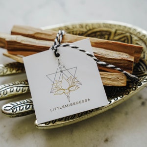 May include: A white tag with a gold geometric lotus flower design and the text "LITTLEMISSDESSA". The tag is tied with twine to a bundle of brown palo santo sticks in a gold metal hand-shaped dish.