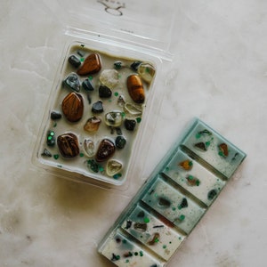 May include: Two wax melts, one in a clear plastic container with a variety of small stones and the other in a rectangular bar with a green and white design.