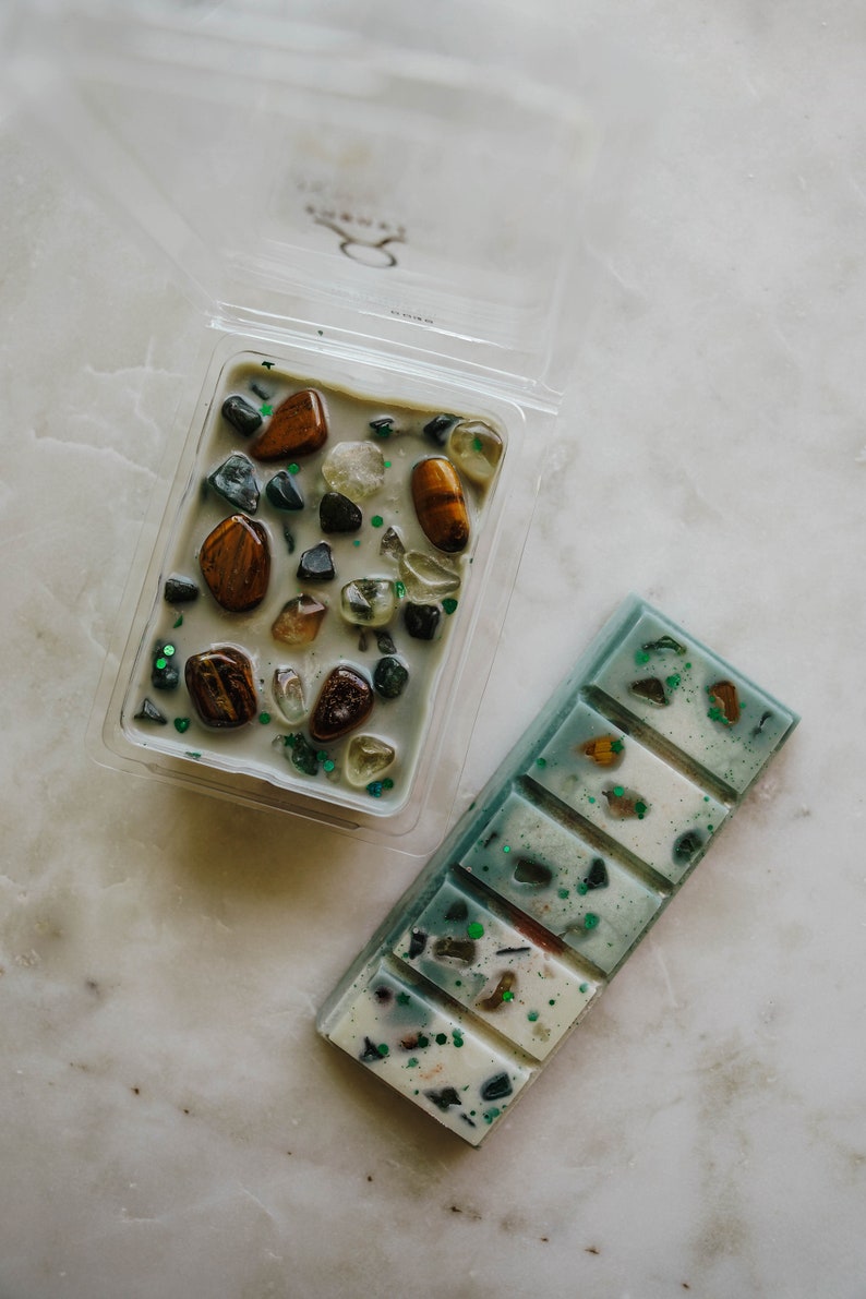 May include: Two wax melts, one in a clear plastic container with a variety of small stones and the other in a rectangular bar with a green and white design.