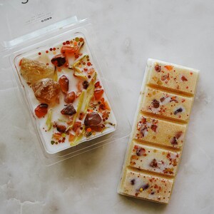 May include: Two wax melts, one in a clear plastic container with a white base and the other in a rectangular bar. The wax melts are white with orange, red, and gold glitter and small pieces of orange and red stones. The container has the text "LEO" and the astrological symbol for Leo.