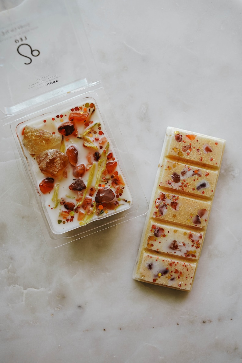 May include: Two wax melts, one in a clear plastic container with a white base and the other in a rectangular bar. The wax melts are white with orange, red, and gold glitter and small pieces of orange and red stones. The container has the text "LEO" and the astrological symbol for Leo.
