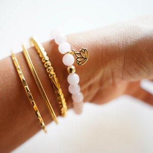 May include: A set of gold bracelets with a white beaded bracelet with a gold lotus charm. The bracelets are stacked on a person's wrist.
