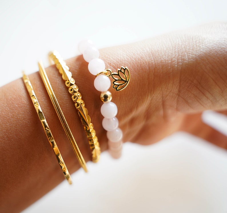 May include: A set of gold bracelets with a white beaded bracelet with a gold lotus charm. The bracelets are stacked on a person's wrist.