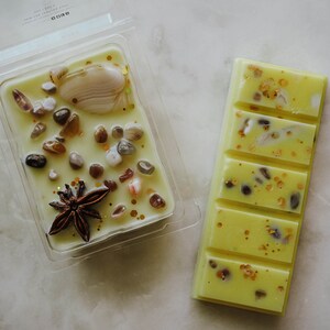 May include: Two wax melts, one in a clear plastic container and one in a bar shape. Both are yellow with embedded glitter and small pieces of seashells. The wax melt in the container also has a star anise.