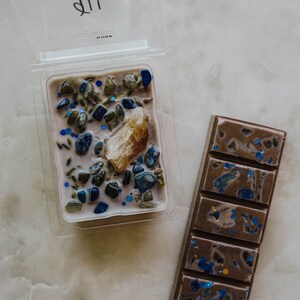 May include: A set of two wax melts, one in a clear plastic container and one in a bar form. The wax melt in the container is a light purple colour with blue and green glitter and small stones. The bar wax melt is a dark brown colour with blue glitter. Both wax melts have the word "VIRGO" printed on the container.