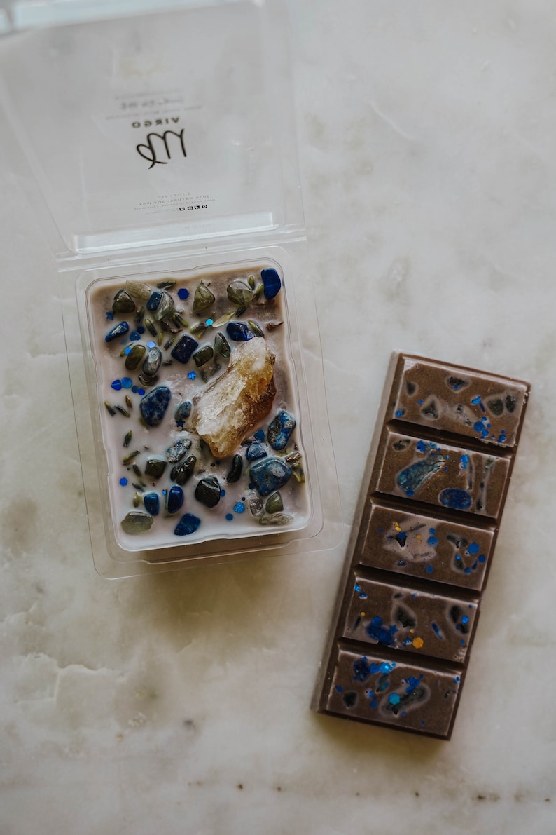 May include: A set of two wax melts, one in a clear plastic container and one in a bar form. The wax melt in the container is a light purple colour with blue and green glitter and small stones. The bar wax melt is a dark brown colour with blue glitter. Both wax melts have the word "VIRGO" printed on the container.