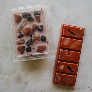 May include: Two wax melts, one in a clear plastic container with a pink wax base and brown and black rock-like pieces, the other a rectangular bar of pink wax with brown and black pieces. The container has the word "ARIES" printed on it.