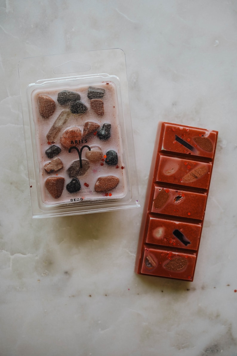May include: Two wax melts, one in a clear plastic container with a pink wax base and brown and black rock-like pieces, the other a rectangular bar of pink wax with brown and black pieces. The container has the word "ARIES" printed on it.