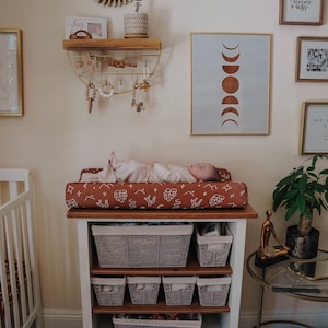May include: A white changing table with three shelves and woven baskets. The changing pad is brown with a white leaf pattern. A baby is lying on the changing pad. The changing table is in a nursery with a crib, a rug, and various wall decorations.
