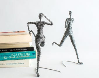 May include: Two silver-colored wire sculptures depicting human figures in motion. One figure is in a running pose, while the other stands with one arm raised. The sculptures are crafted from thin, coiled metal wire, creating a textured, semi-transparent appearance for each form.