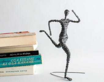 May include: A sculpture of a human figure in a running pose, constructed from thin, dark grey metallic wire wrapped in a dense, textured pattern. The figure is balanced on a single wire base, with one leg extended forward and arms bent.