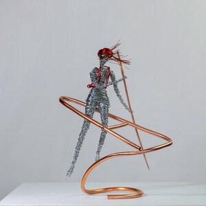 Handmade female rafter wire sculpture holding a paddle, with red hair, a copper wire raft and aluminum wire details.