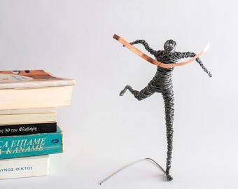 May include: A sculpture of a human figure made from coiled silver-toned wire, depicted in a running pose. The figure is crossing a finish line represented by a flat, horizontal copper-colored ribbon held across the chest and outstretched arms.
