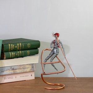 Handmade female rafter wire sculpture holding a paddle, with red hair, a copper wire raft and aluminum wire details.