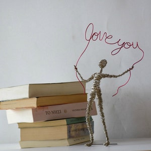 May include: A silver wire sculpture of a person holding a red wire sculpture that says "love you". The sculpture is standing on a stack of books.