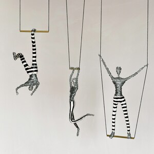 K&ouml;nnte beinhalten: Drei Drahtfiguren, jede mit einer schwarzen und wei&szlig;en gestreiften Hose, h&auml;ngen an Schn&uuml;ren von der Decke. Jede Figur h&auml;lt eine horizontale Stange fest. Die Figuren sind in verschiedenen Positionen dargestellt, eine h&auml;ngt kopf&uuml;ber, eine steht gerade und eine ist in einer Position in der Luft.