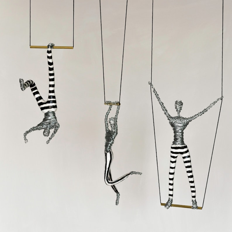 K&ouml;nnte beinhalten: Drei Drahtfiguren, jede mit einer schwarzen und wei&szlig;en gestreiften Hose, h&auml;ngen an Schn&uuml;ren von der Decke. Jede Figur h&auml;lt eine horizontale Stange fest. Die Figuren sind in verschiedenen Positionen dargestellt, eine h&auml;ngt kopf&uuml;ber, eine steht gerade und eine ist in einer Position in der Luft.
