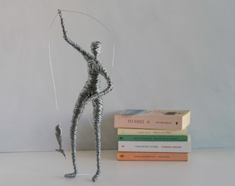 May include: A silver-colored wire sculpture of a person standing and holding a fishing rod with a small wire fish attached to the line. The sculpture is constructed from coiled metal wire and is positioned next to a stack of five books.