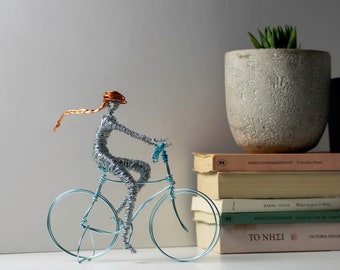 May include: A wire sculpture of a person riding a bicycle. The bicycle is constructed from light blue wire, while the person is made of silver wire with a copper-colored wire head and a long, flowing ponytail extending behind the figure.