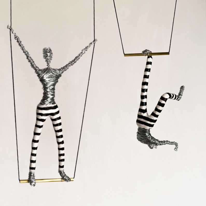 K&ouml;nnte beinhalten: Zwei Drahtfiguren von Menschen in schwarz-wei&szlig; gestreiften Hosen, die an Schn&uuml;ren von der Decke h&auml;ngen. Die Figuren halten eine goldene Stange.