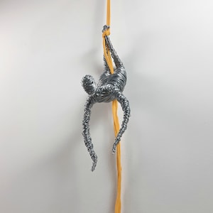 Può includere: Una scultura in filo d'argento di una persona che scala una corda gialla. La scultura è sospesa dal soffitto e sembra arrampicarsi verso l'alto.