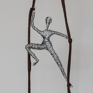 May include: A silver wire sculpture of a person climbing a brown rope. The sculpture is suspended from the ceiling.