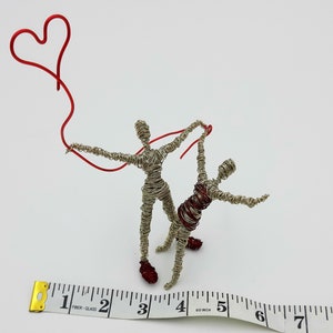 Può includere: Scultura in filo metallico di due figure che tengono un palloncino rosso a forma di cuore. Le figure sono in filo d'argento e sono in piedi su una superficie bianca. Un metro a nastro è in primo piano.
