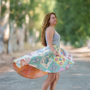 May include: A woman wearing a light blue tank top and a colorful patchwork skirt with floral designs. The skirt is flowing in the wind.