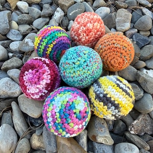 May include: A collection of seven spherical crochet balls featuring various color patterns including rainbow stripes, solid orange, variegated teal, pink gradients, and yellow, gray, and white diagonal stripes, all crafted with a textured yarn stitch.