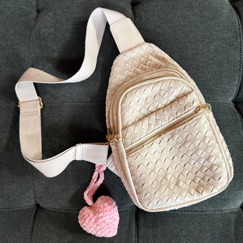 K&ouml;nnte beinhalten: Cremefarbene, gewebte Umh&auml;ngetasche mit goldenem Rei&szlig;verschluss und wei&szlig;em Riemen. Ein kleiner, rosa geh&auml;kelter Herzanh&auml;nger h&auml;ngt an der Tasche. Die Tasche hat eine Vordertasche und ein Hauptfach.
