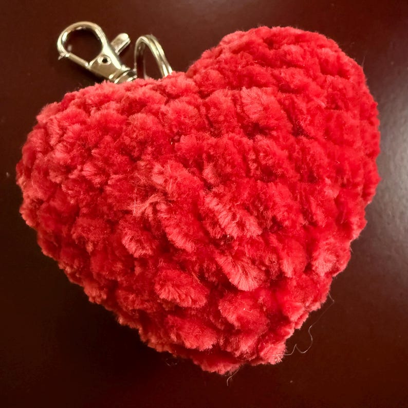 K&ouml;nnte beinhalten: Ein roter, herzf&ouml;rmiger Schl&uuml;sselanh&auml;nger aus Pl&uuml;sch mit einem silberfarbenen Verschluss. Das Herz hat eine strukturierte, flauschige Oberfl&auml;che. Ein dekoratives Accessoire.