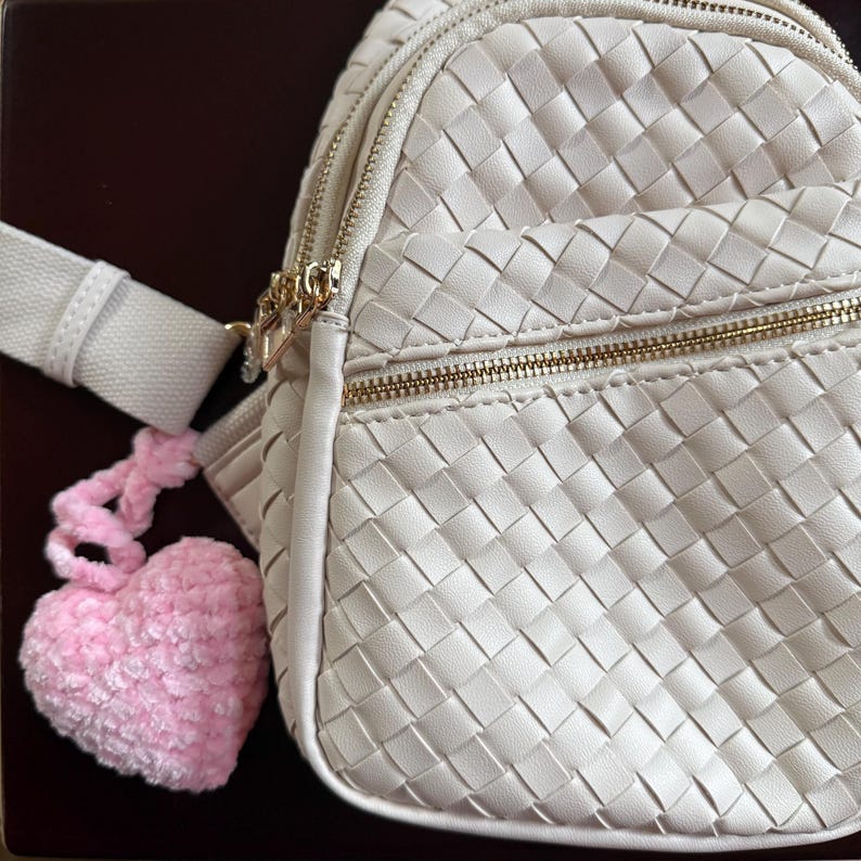 K&ouml;nnte beinhalten: Cremefarbener Rucksack mit Webmuster, goldfarbenen Rei&szlig;verschl&uuml;ssen und passendem Riemen. Ein flauschiger, herzf&ouml;rmiger rosa Anh&auml;nger h&auml;ngt am Rei&szlig;verschluss. Die Tasche hat ein strukturiertes, korb&auml;hnliches Design.