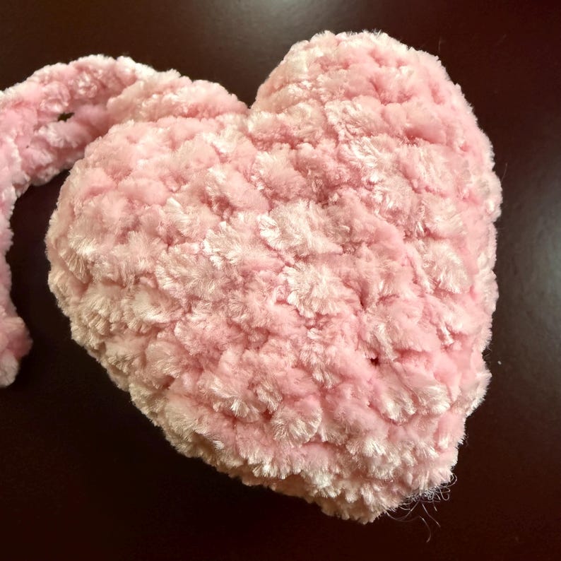 K&ouml;nnte beinhalten: Ein herzf&ouml;rmiges Kissen in Rosa mit einer weichen, strukturierten Oberfl&auml;che. Das Kissen besteht aus einem flauschigen Material und hat einen hellrosa Farbton. Das Bild zeigt eine Nahaufnahme des Herzens und hebt seine flauschige Textur hervor.