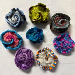 May include: A collection of nine handmade crochet coasters in various colors. Each coaster features a spiral design with a ruffled edge. Colors include blue, green, pink, purple, and white, offering a vibrant and artistic touch to any home decor.