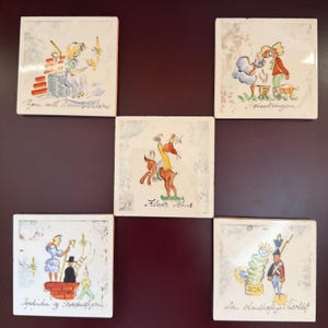 May include: Five square ceramic tiles featuring hand-painted illustrations of fairy tale characters. Each tile displays a unique scene with handwritten text in Danish, including Pigen med Svovlstikkerne, Svinedrengen, Klods Hans, Hyrdinden og Skorstensfejeren, and Den standhaftige tinsoldat.